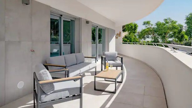 A modern balcony with a sofa, armchair, and coffee table, featuring large windows and a view of greenery.
