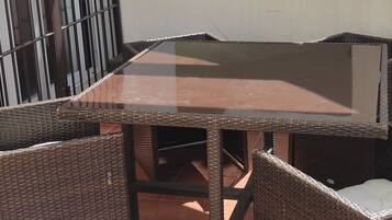 Un patio con mesa y sillas de mimbre, suelo de baldosas y vistas a un edificio con ventanas y puerta.