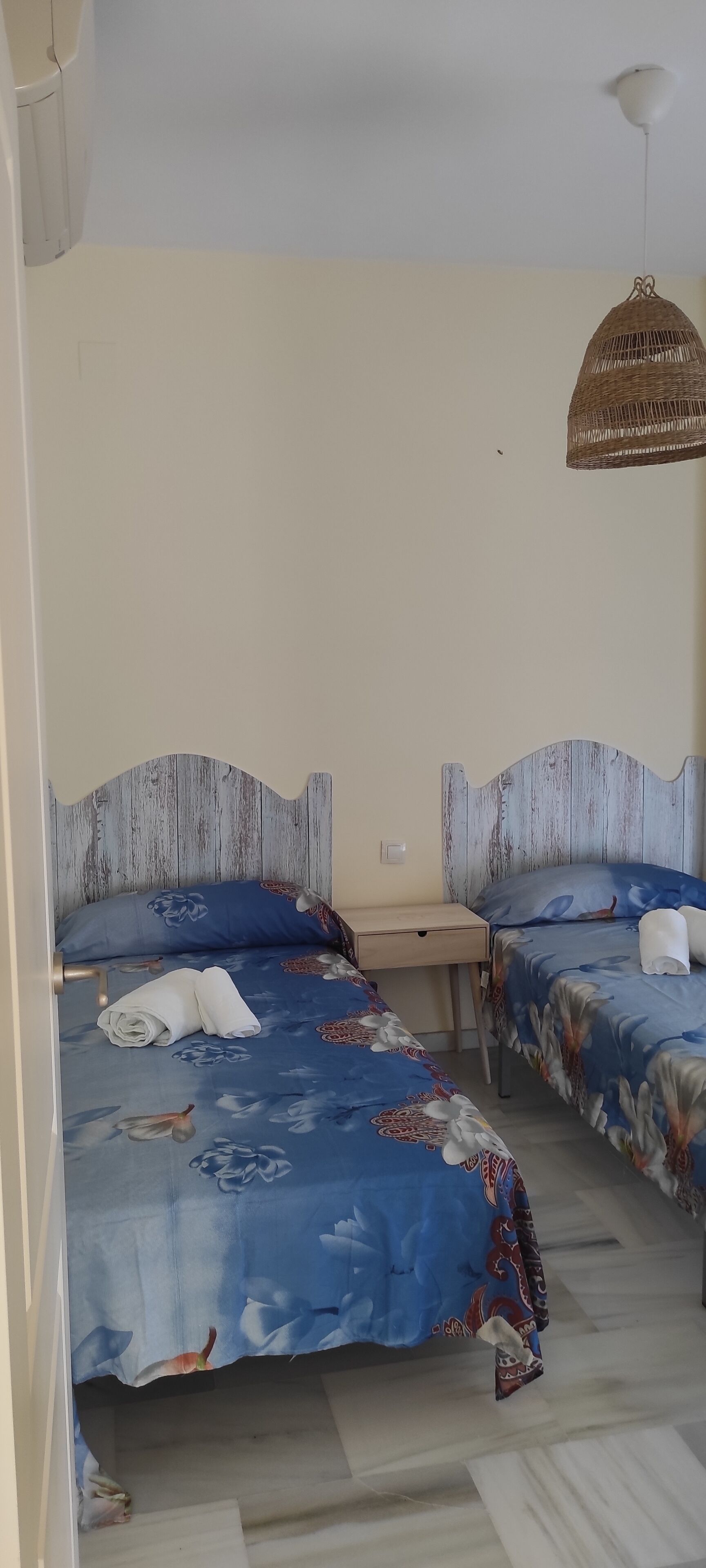 A room with two beds, each with a blue floral bedspread and white pillows. There is a wicker hanging light fixture above the beds.