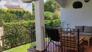 Un balcon avec une table en bois, un canapé et une chaise. Il y a un jardin avec une clôture et diverses plantes.