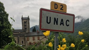 Una señalización con 'D2' y 'UNAC' en una carretera rural con una iglesia y montañas al fondo.