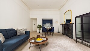 Een woonkamer met een blauwe bank, een bijzettafel met fruit, een open haard en een eethoek met stoelen en een tafel.