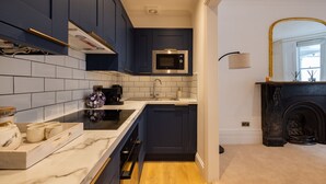 Een kleine keuken met donkerblauwe kastjes, een wit marmeren aanrecht, een ingebouwde magnetron en een zwarte open haard.
