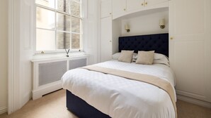 Een slaapkamer met een groot raam, witte wanden, een donker hoofdeinde en een beige sprei.