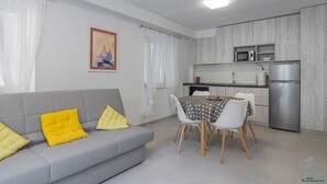 Un saló modern amb un sofà gris, coixinets grocs i una taula de menjador amb cadires blanques. Es veu una cuina amb microones i nevera al fons.