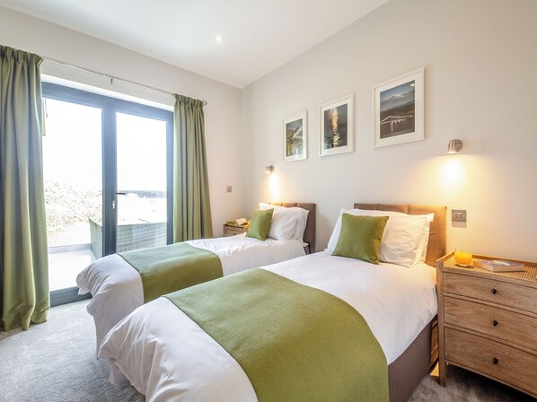 Habitación de hotel con dos camas, un ventanal grande con cortinas, una mesita de noche de madera y tres cuadros enmarcados en la pared.