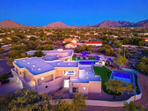 Un immeuble moderne à plusieurs étages, doté d’une piscine sur le toit et d’un court de tennis, entouré d’une végétation désertique et de montagnes.