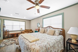 A bedroom with a large bed, a ceiling fan, two windows with blinds, a bedside table with a lamp, and a bench in the corner.