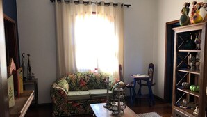 Un salon avec un canapé à motifs floraux, une table en bois sur laquelle se trouve une lampe, une petite table ronde et un meuble en bois orné d’objets décoratifs.