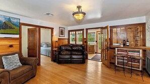 A spacious living room with a leather sofa, wooden flooring, a bar area with stools, and a large TV mounted on the wall. - Bearpaw Bungalow | Fenced Yard · Near Lake (Big Bear Lake)