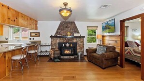 A cozy living room with a fireplace, a bar area with stools, and a comfortable seating area with a sofa and a television. - Bearpaw Bungalow | Fenced Yard · Near Lake (Big Bear Lake)