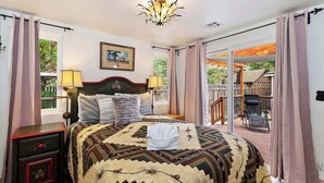 A bedroom with a large bed, a nightstand, a chandelier, a window with curtains, and a balcony with a chair. - Bearpaw Bungalow | Fenced Yard · Near Lake (Big Bear Lake)