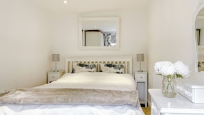 A neatly arranged bedroom with a bed, bedside tables, lamps, a mirror, and a vase with flowers.