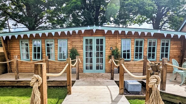 A wooden cabin with a front porch, a rope barrier, and a small outdoor seating area.