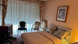Une chambre d’hôtel avec un lit, un bureau, deux chaises, un radiateur et une fenêtre avec des rideaux.