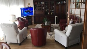 Un salon comprenant un téléviseur, deux fauteuils, un pouf rouge, une table basse et une bibliothèque.