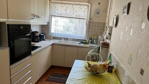 Une petite cuisine équipée d’une cuisinière, d’un évier et d’une fenêtre. On y trouve une table avec un panier de pain et une bouteille de lait.