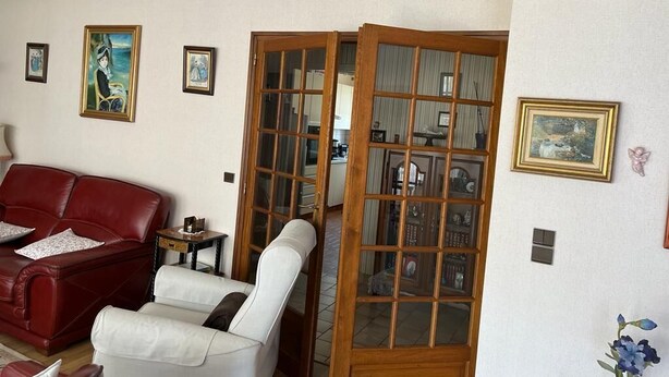 Un salon avec un canapé rouge, un fauteuil blanc et une porte-moustiquaire en bois donnant sur un coin cuisine.