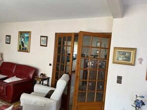 A living room with a red sofa, a white armchair, and a wooden screen door leading to a kitchen area.