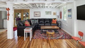 A modern living room with a black leather sectional sofa, a small white table, a wooden coffee table, and a colorful rug. - Folly Vacation 305 Beach Home (Folly Beach)