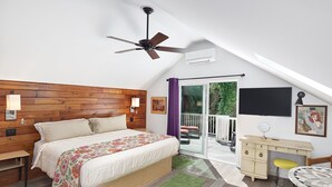 A bedroom with a wooden headboard, a bed with a floral bedspread, a green rug, a desk with a mirror, and a TV on the wall. - Folly Vacation 305 Beach Home (Folly Beach)