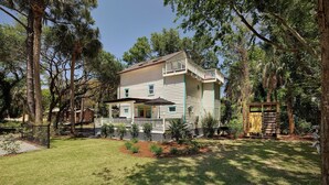 A two-story house with a porch and a small deck, surrounded by trees and a well-maintained lawn. - Folly Vacation 305 Beach Home (Folly Beach)