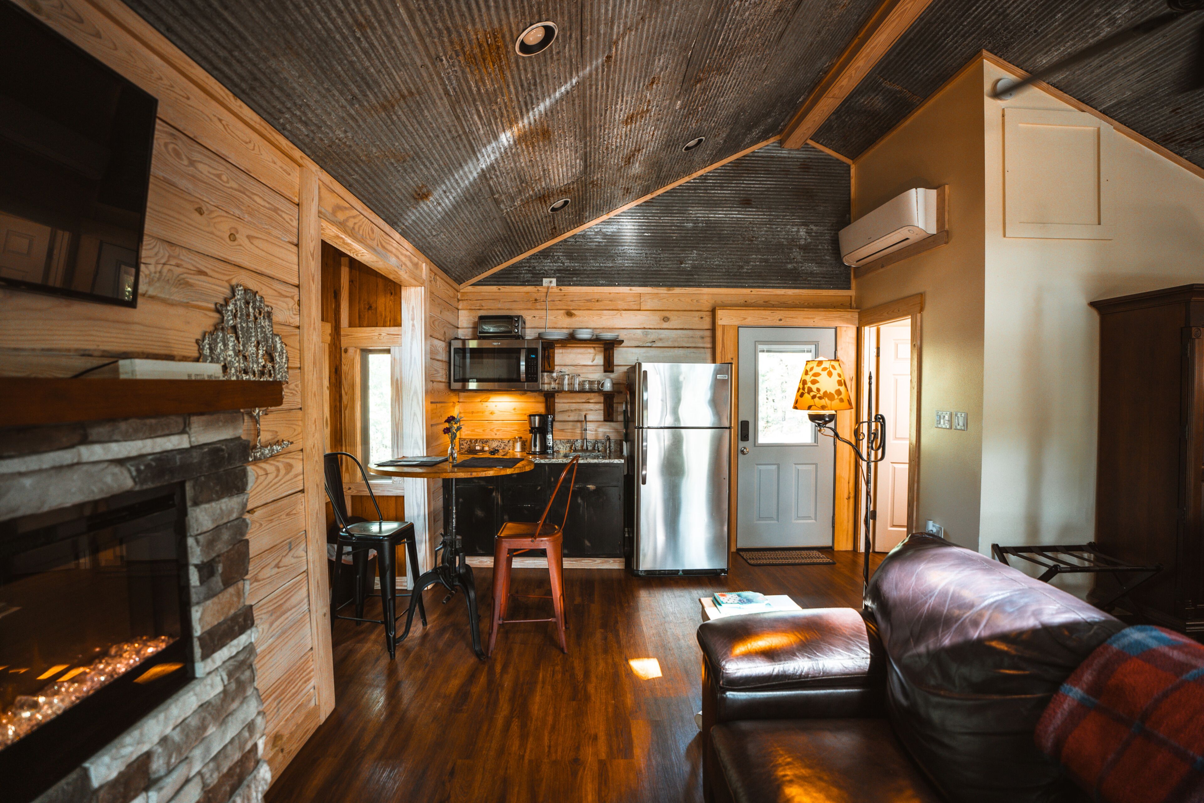 A cozy cabin interior with a stone fireplace, wooden walls, a dining area with a table and chairs, a kitchen with modern appliances, and a leather sofa.