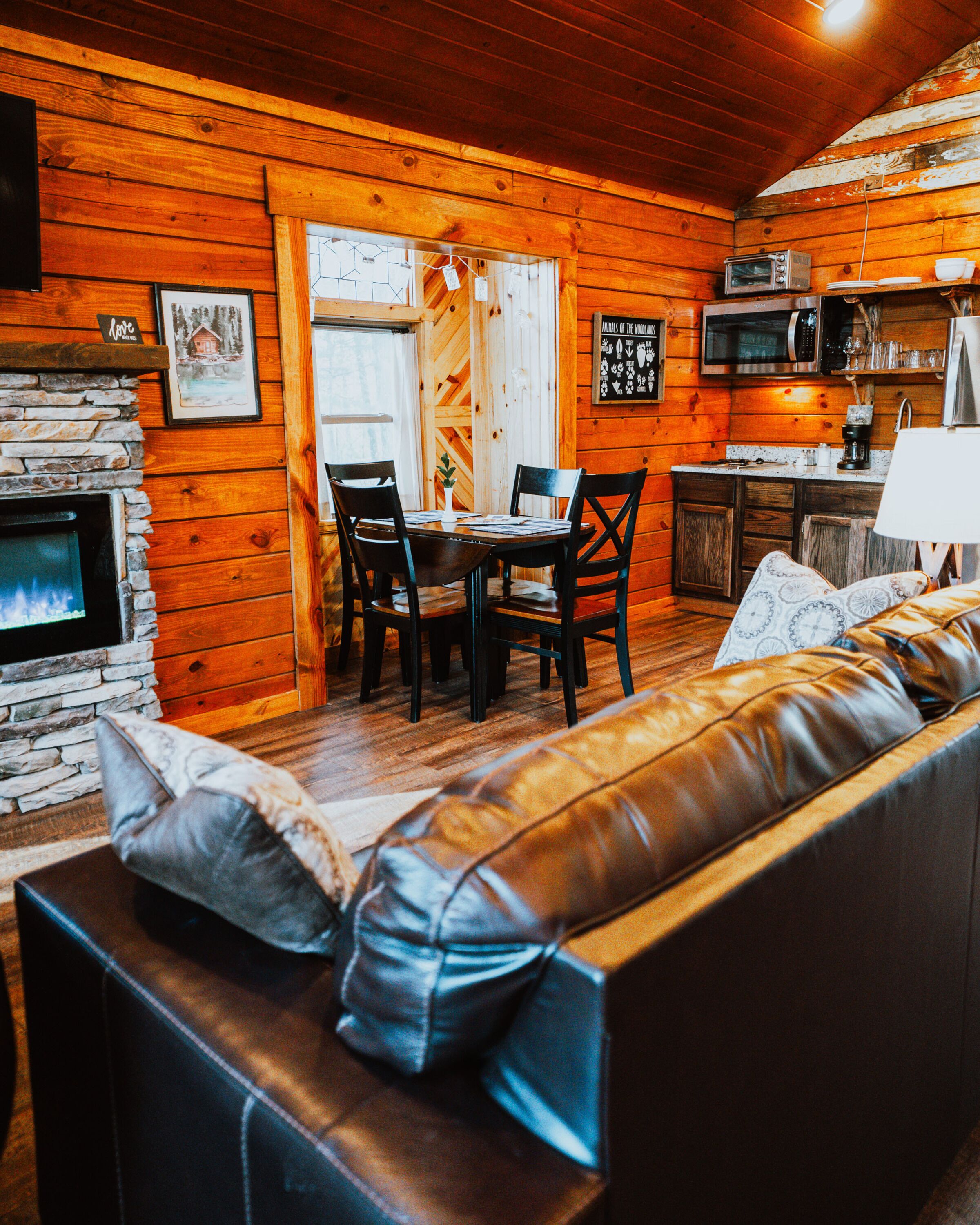 A cozy cabin interior with a stone fireplace, wooden walls, a leather sofa, a dining table with chairs, and a kitchen area with a microwave.