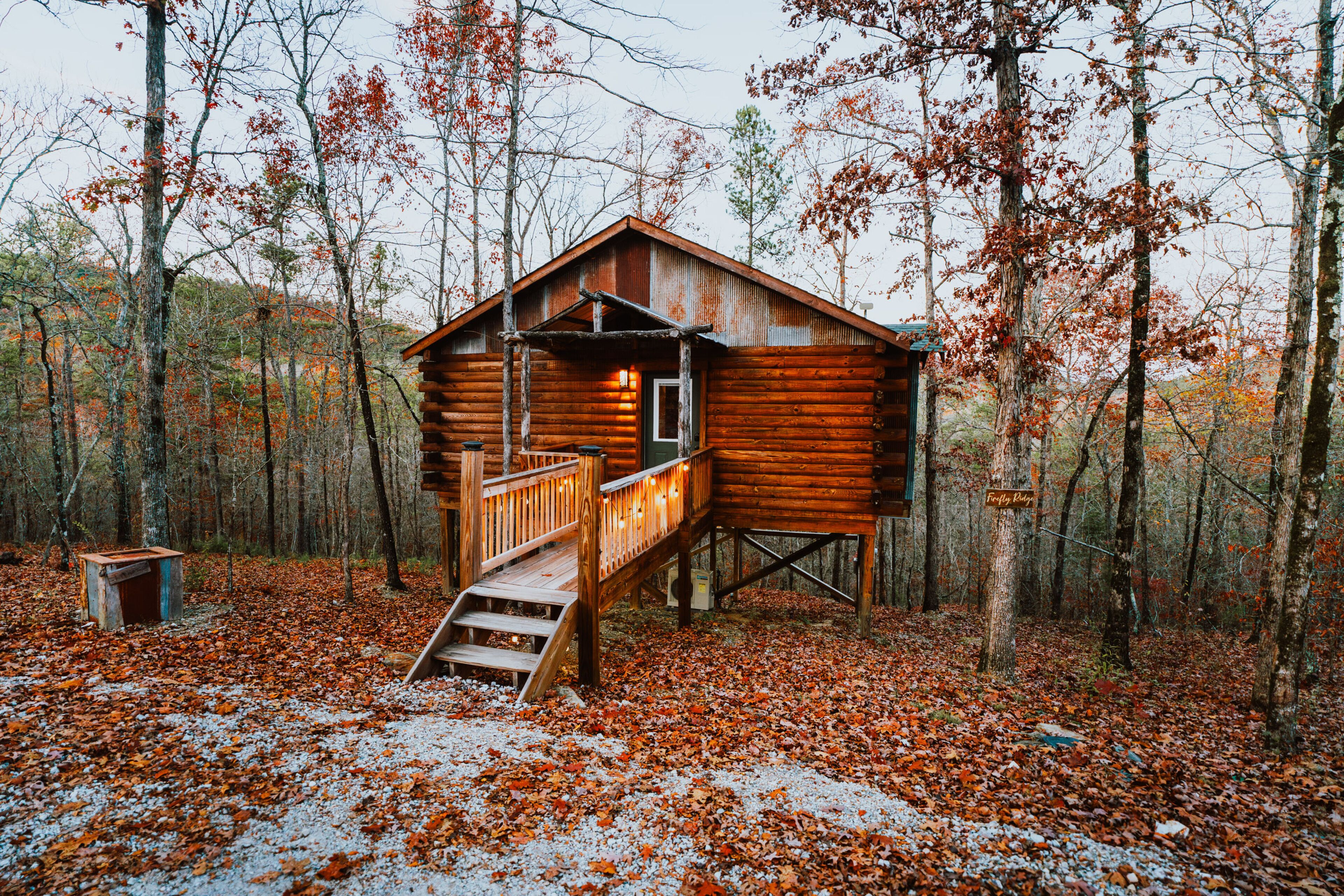 A wooden cabin with a wooden staircase leading to a deck, surrounded by a forest with autumn leaves.