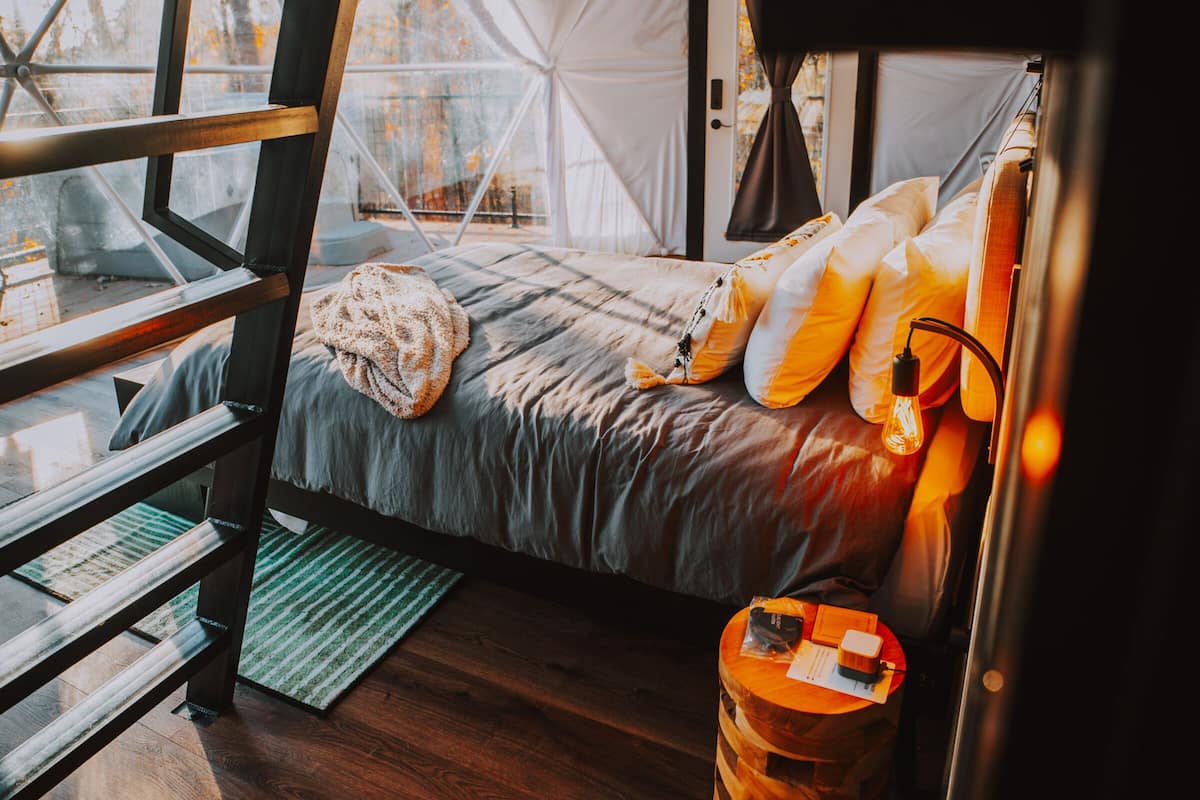 A cozy, modern tent-style bedroom with a bed, pillows, a small wooden table, and a view of the outdoors.
