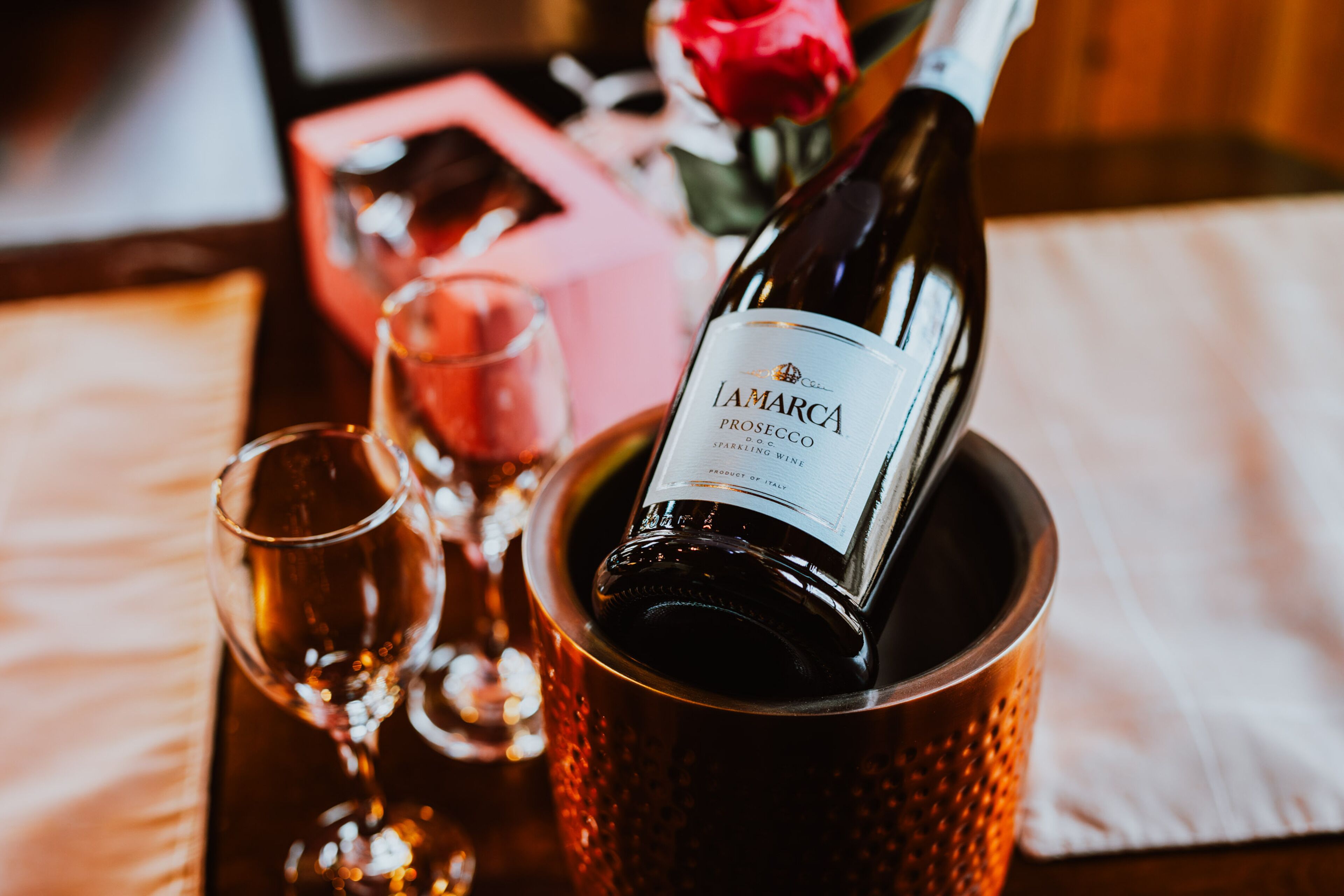 Una botella de vino espumoso Lamarca Prosecco en un cubo de cobre con copas y una rosa.