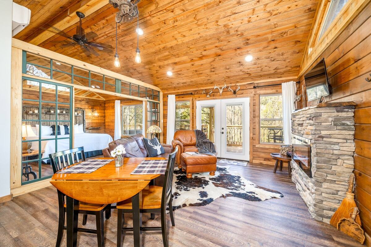A spacious room with a wooden ceiling, a dining area with a table and chairs, a leather sofa, and a stone fireplace.