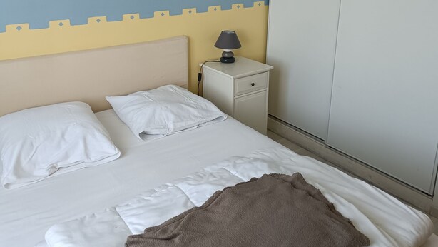A neatly made bed with white linens and a brown throw blanket, a bedside lamp, and a wall-mounted light fixture. - Beautiful house near the ocean—peaceful, charming, and perfect for a family vacation (Meschers-sur-Gironde)