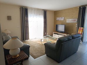 A living room with a sofa, a glass coffee table, a TV on a wooden stand, and a window with curtains.