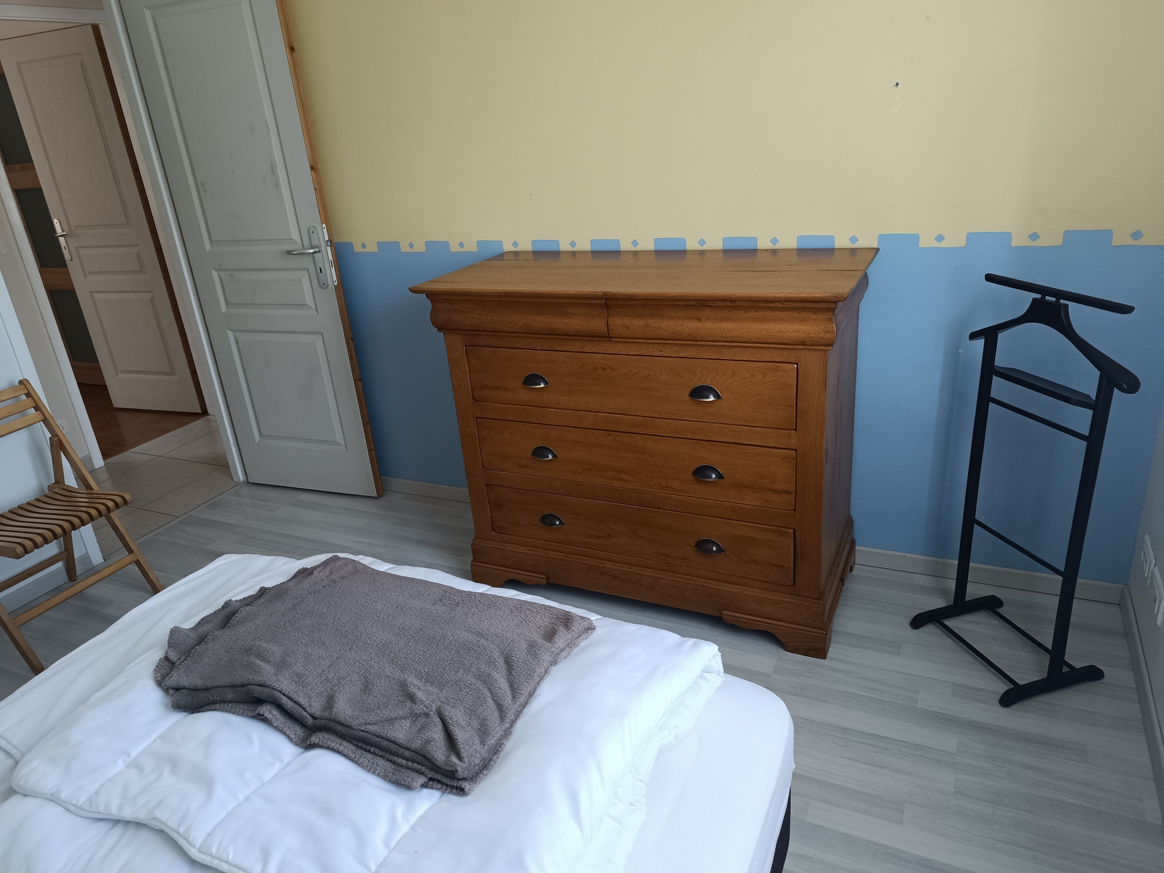 Une chambre à coucher avec un lit, une commode en bois, une chaise et un portant à vêtements.
