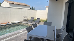 Ein Poolbereich mit Tisch und Stühlen, einem gefliesten Pool und einer weißen Wand mit einer gepflasterten Dachkonstruktion.