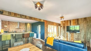 A modern living room with a blue sofa, wooden dining table, and a kitchen area with a brick wall and wooden paneling.