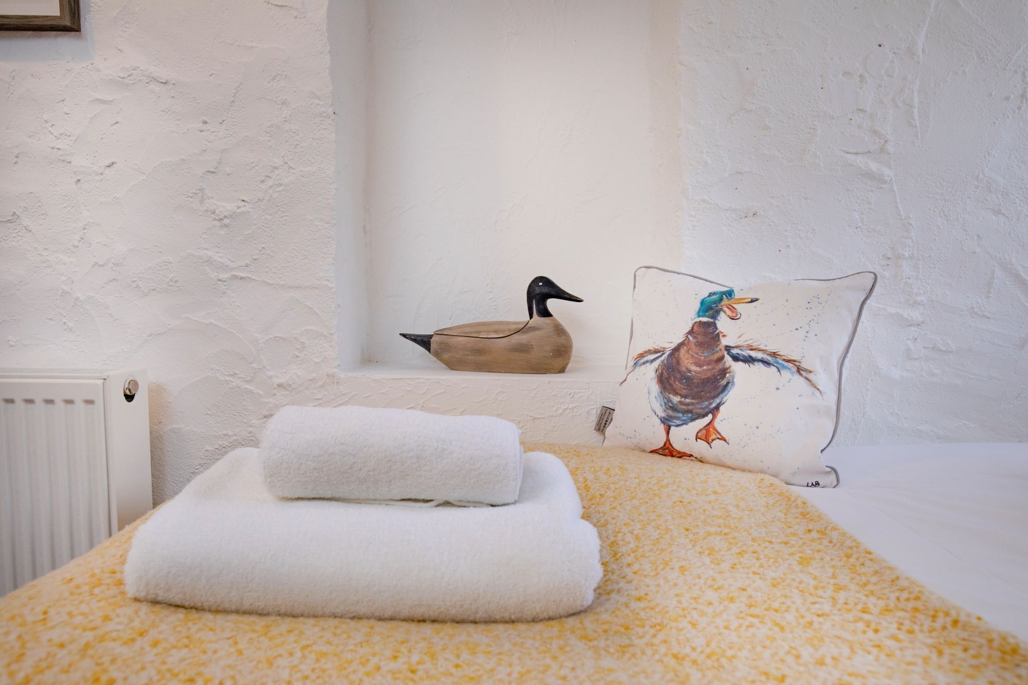 A neatly made bed with a yellow patterned bedspread, a white towel, and a pillow with a bird design.
