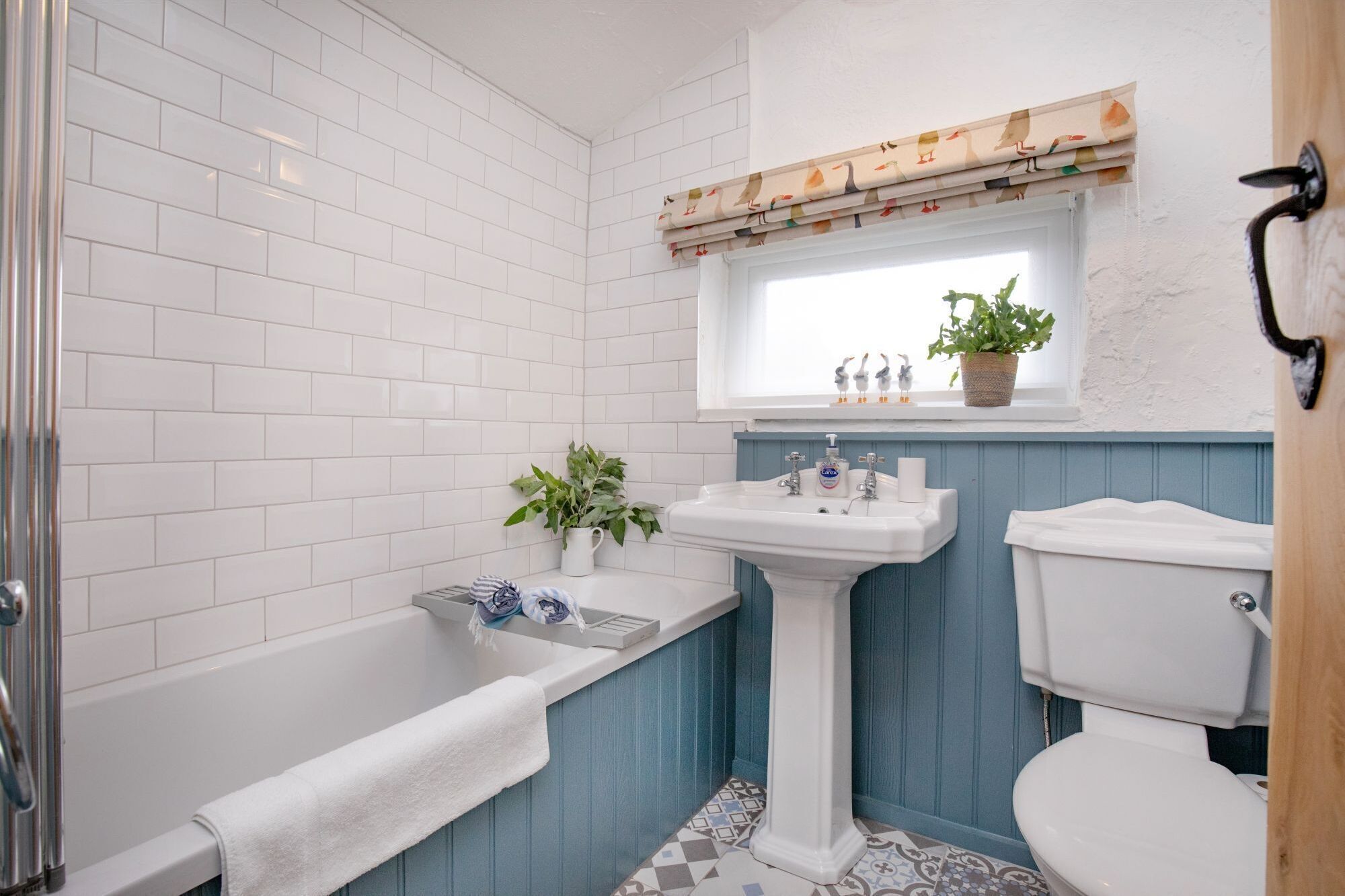 Ein Badezimmer mit weißen Fliesen, einer Badewanne, einem Standwaschbecken und einem Fenster mit Vorhang.