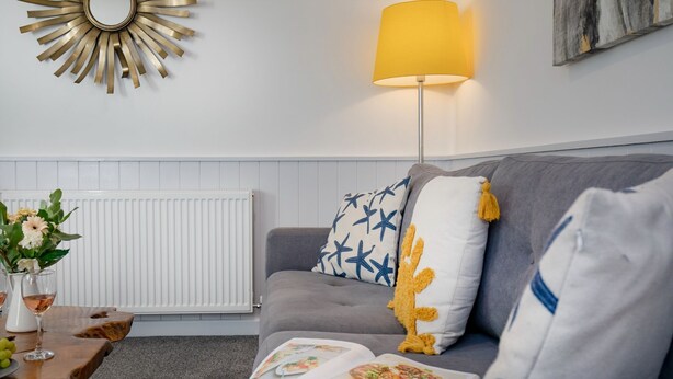 A living room with a grey sofa, a yellow floor lamp, a white radiator, and a decorative sunburst mirror. - Salty Dog Apartment @ Sunnybeach (Paignton)