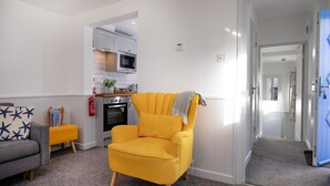 A living room with a yellow armchair, a grey sofa, and a small side table with a plant. A kitchen is visible in the background with white cabinets and a microwave. - Salty Dog Apartment @ Sunnybeach (Paignton)