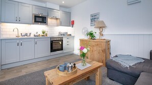 A modern kitchen with white cabinets, a microwave, and an oven. There's a wooden table with a vase of flowers and a tray with mugs. A grey sofa and a lamp are in the living area. - Anchors Away Apartment @ Sunnybeach (Paignton)