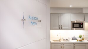 A modern kitchen with white cabinets, a stainless steel microwave, and a sink. A sign on the wall reads "Anchors Away". - Anchors Away Apartment @ Sunnybeach (Paignton)