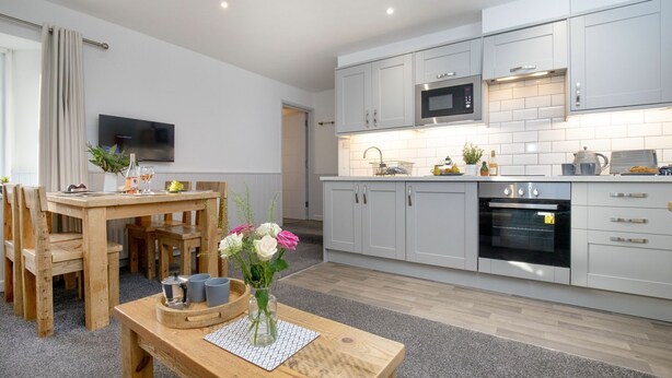 A modern kitchen with white cabinets, a microwave, and an oven. There is a dining area with a wooden table and chairs, and a small vase with flowers on the table. - Anchors Away Apartment @ Sunnybeach (Paignton)