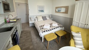 Kompakti hotellihuone, jossa on pieni keittiönurkkaus, kuviollinen päiväpeite, yöpöytä lampun kanssa ja keltainen nojatuoli.