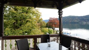 A covered outdoor seating area with a table and chairs, overlooking a lake with trees and hills in the background.