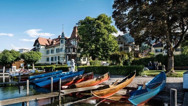 A calm waterway with several wooden boats docked at a wooden pier. In the background, there are buildings with traditional architecture and a group of people gathered on the grass.