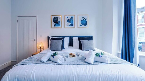 Ein ordentlich bezogenes Bett mit einer blau-weißen Tagesdecke, ein Kopfteil mit dunklen und hellen Kissen und drei gerahmte abstrakte Kunstwerke an der Wand.