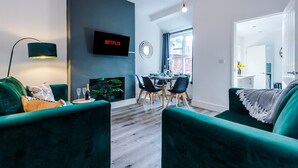 Ein modernes Wohnzimmer mit einem grünen Sofa, einem Essbereich mit Tisch und Stühlen, einem flachbildfernseher an der Wand und einem großen Fenster, das für natürliches Licht sorgt.