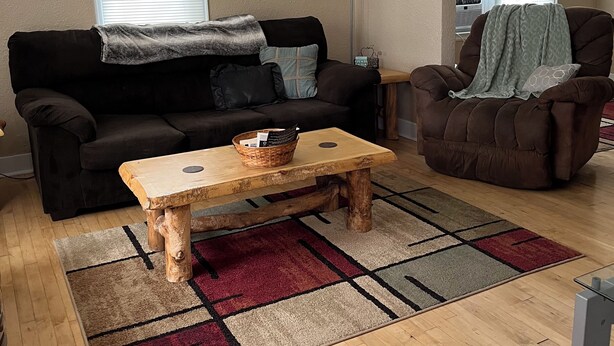 Ein Wohnzimmer mit Couchtisch aus Holz, einer schwarzen Couch, einem braunen Sessel und einem gemusterten Teppich.